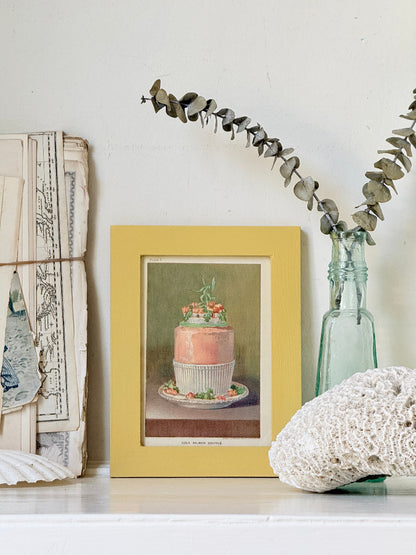 Framed Victorian cookery illustration of a celebration cake from 1896 in a mustard yellow frame displayed on a white surface.