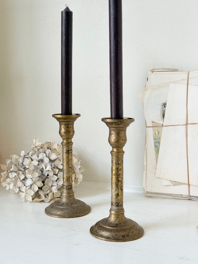 Pair of tall Indian engraved brass candlesticks in the Benares style with floral detailing and black taper candles on a white surface