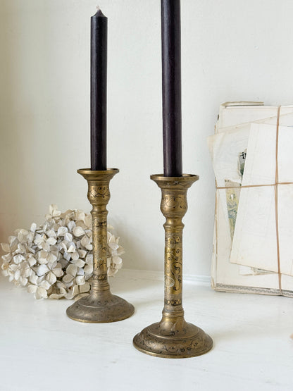 Pair of tall Indian engraved brass candlesticks in the Benares style with floral detailing and black taper candles on a white surface