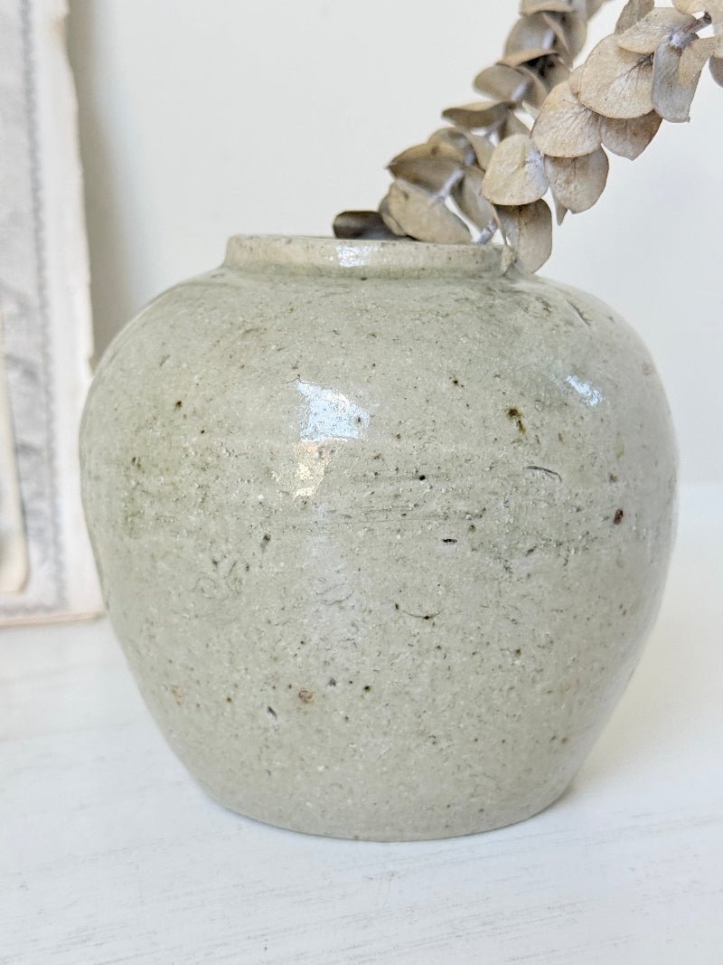 A small round ceramic vase with a speckled light gray-green glaze, holding dried beige flowers, set against a soft white background.