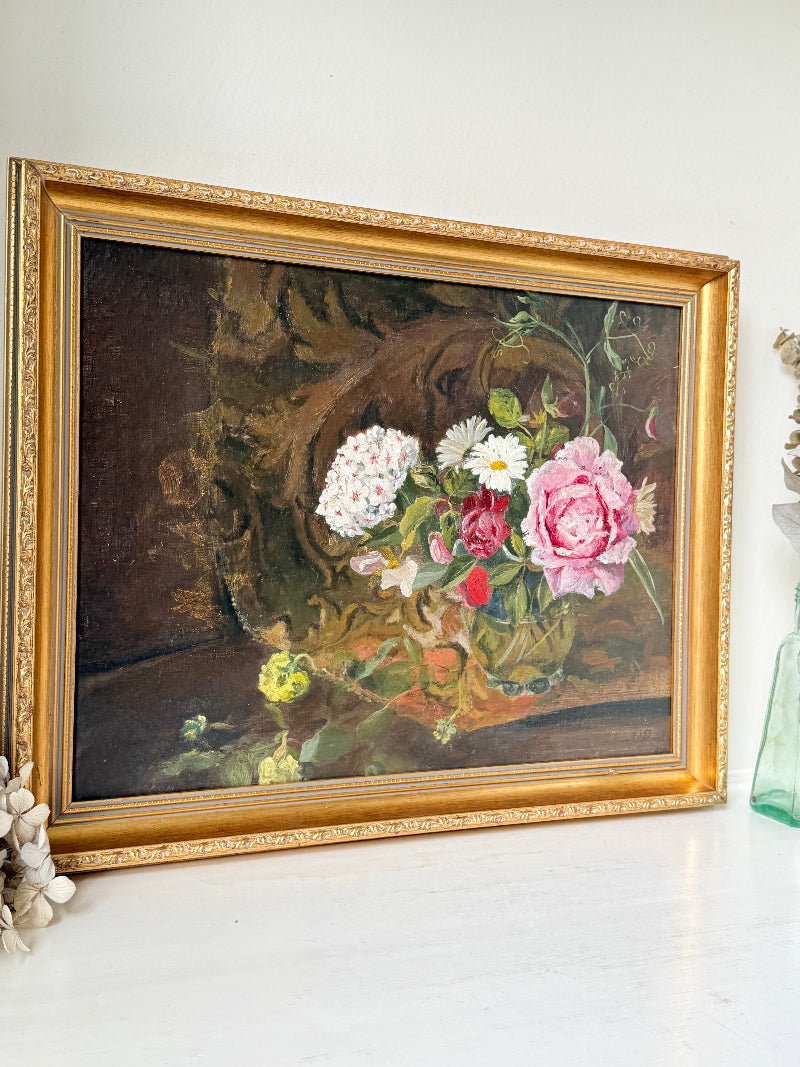A framed oil painting of a still life floral arrangement, featuring pink and white roses with green leaves, housed in an ornate gold frame and displayed on a white surface against a white wall.