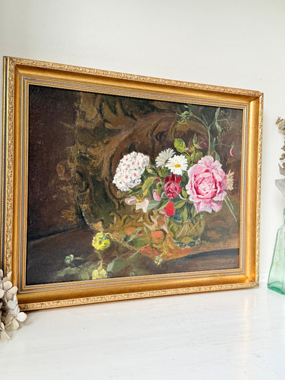 A framed oil painting of a still life floral arrangement, featuring pink and white roses with green leaves, housed in an ornate gold frame and displayed on a white surface against a white wall.