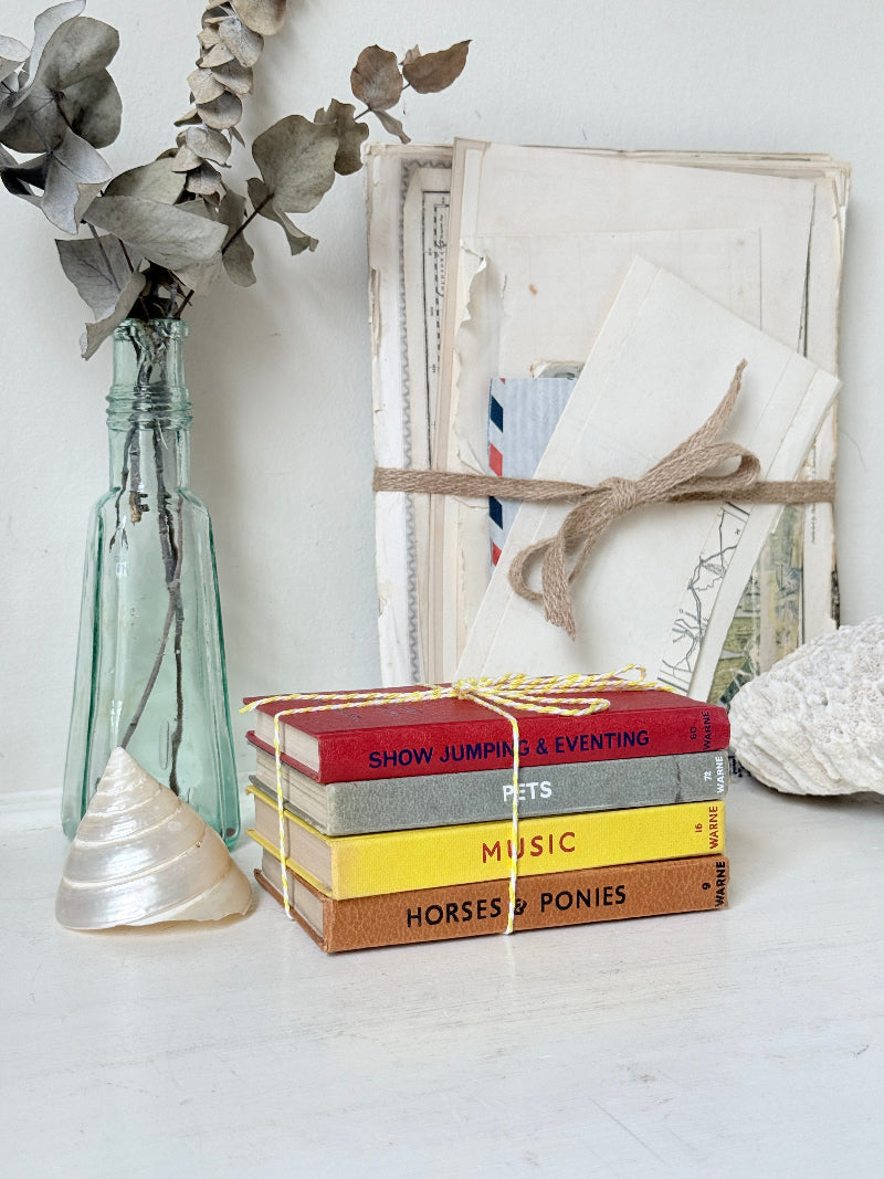 A styled flat-lay featuring a stack of colorful hardcover books tied with yellow string, set against a white background with decorative props including a glass bottle with dried leaves, a shell, and a wrapped bundle of papers.