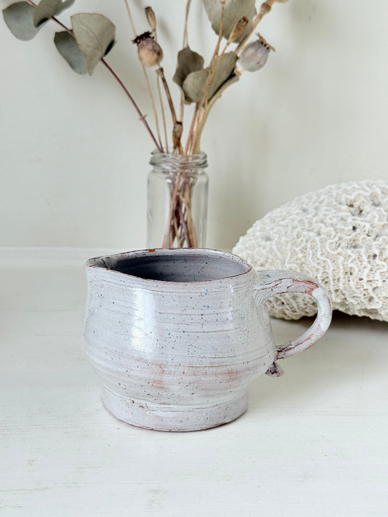 Studio pottery milk jug with soft pink and grey speckled glaze, photographed on a white surface