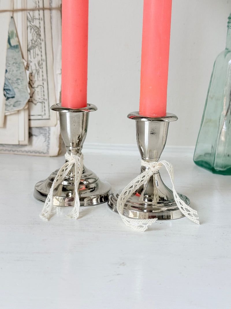 Pair of silver tone candlesticks with urn sconces and round stepped bases holding coral candles, photographed on a white surface