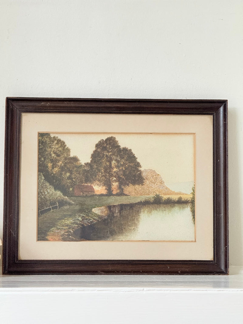 Antique framed rural watercolour of river, trees, and red-roofed building, in dark wooden frame with cream mount, on white surface.