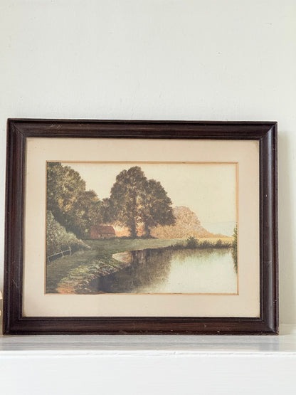 Antique framed rural watercolour of river, trees, and red-roofed building, in dark wooden frame with cream mount, on white surface.