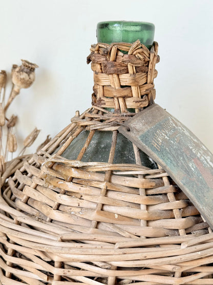 antique French demi john olive oil jar in wicker basket with green glass bottle and painted metal label, displayed on white surface