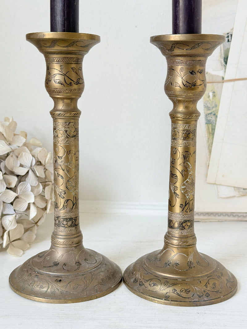 Pair of tall Indian engraved brass candlesticks in the Benares style with floral detailing and black taper candles on a white surface