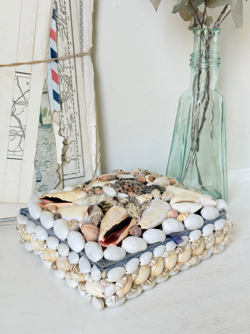 A square decorative pillow or cushion covered with white, beige, and brown seashells and small stones, styled on a white surface next to a tall translucent green glass bottle vase.