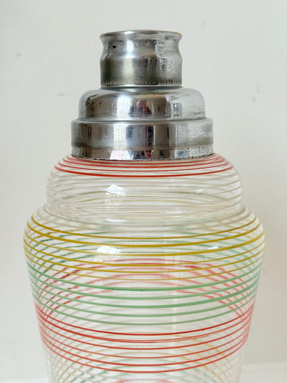Vintage glass cocktail shaker with multicoloured stripes and silver lid photographed on a white surface.