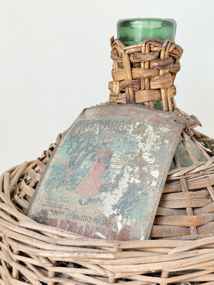 antique French demi john olive oil jar in wicker basket with green glass bottle and painted metal label, displayed on white surface