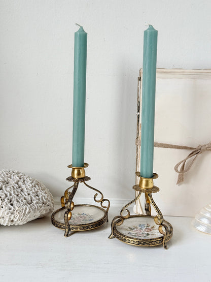 A pair of vintage-style candlestick holders with ornate gold-toned metal frames and floral porcelain bases, holding tall sage green taper candles, styled on a white surface against a white wall.
