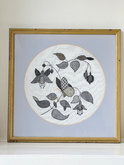 Framed circular hand embroidery of birds and fuchsia flowers in black and grey on white linen, set in gold frame, white surface background.
