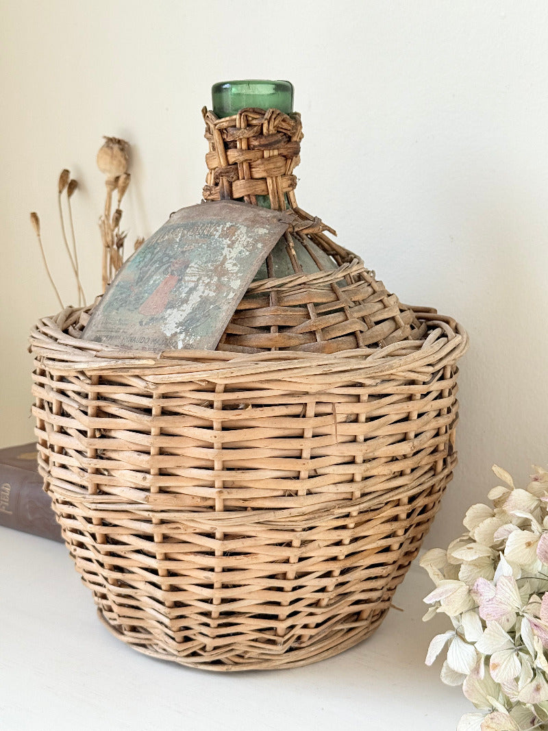 antique French demi john olive oil jar in wicker basket with green glass bottle and painted metal label, displayed on white surface