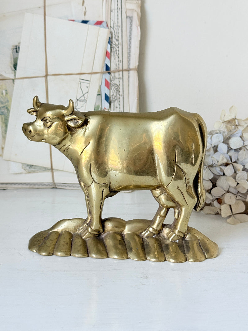 A polished brass cow figurine standing on a decorative base, displayed on a white surface with a softly blurred background of vintage letters and dried flowers.