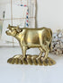 A polished brass cow figurine standing on a decorative base, displayed on a white surface with a softly blurred background of vintage letters and dried flowers.