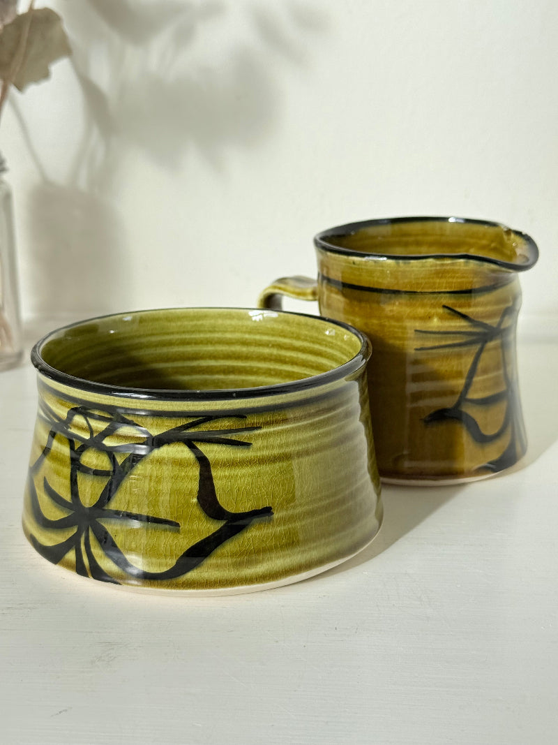 Studio pottery sugar bowl and milk jug in olive green glaze with black brushstroke decoration, photographed on a white surface.