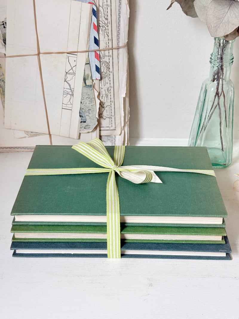 A stack of green hardcover books or albums tied with a pale green and white striped ribbon, set on a white surface with vintage papers and a glass bottle in the background.