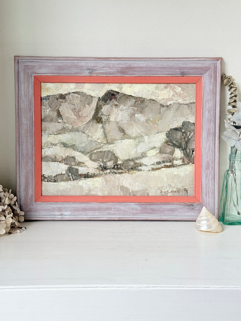 A framed landscape painting with textured brushstrokes in earthy beige, gray, and brown tones, set in a distressed lavender-gray frame with a coral inner border, displayed on a white surface with decorative seashells and a glass vase.