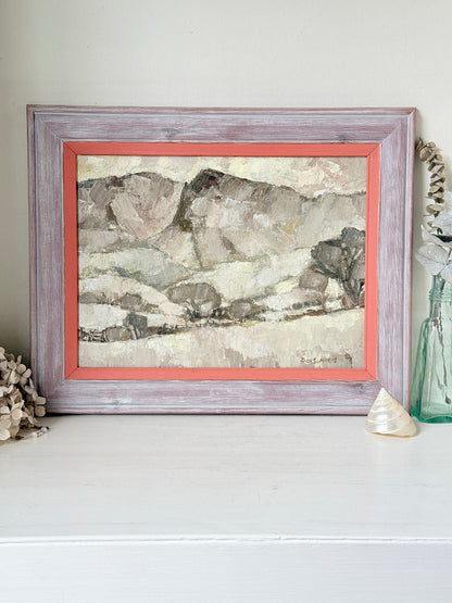 A framed landscape painting with textured brushstrokes in earthy beige, gray, and brown tones, set in a distressed lavender-gray frame with a coral inner border, displayed on a white surface with decorative seashells and a glass vase.