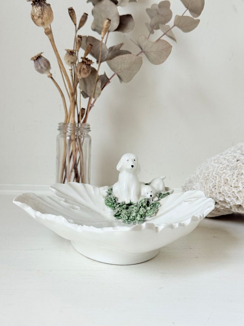 Italian ceramic dish featuring two white dogs on a sculpted leaf base, photographed on a white surface.