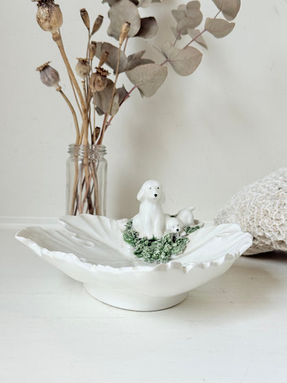 Italian ceramic dish featuring two white dogs on a sculpted leaf base, photographed on a white surface.