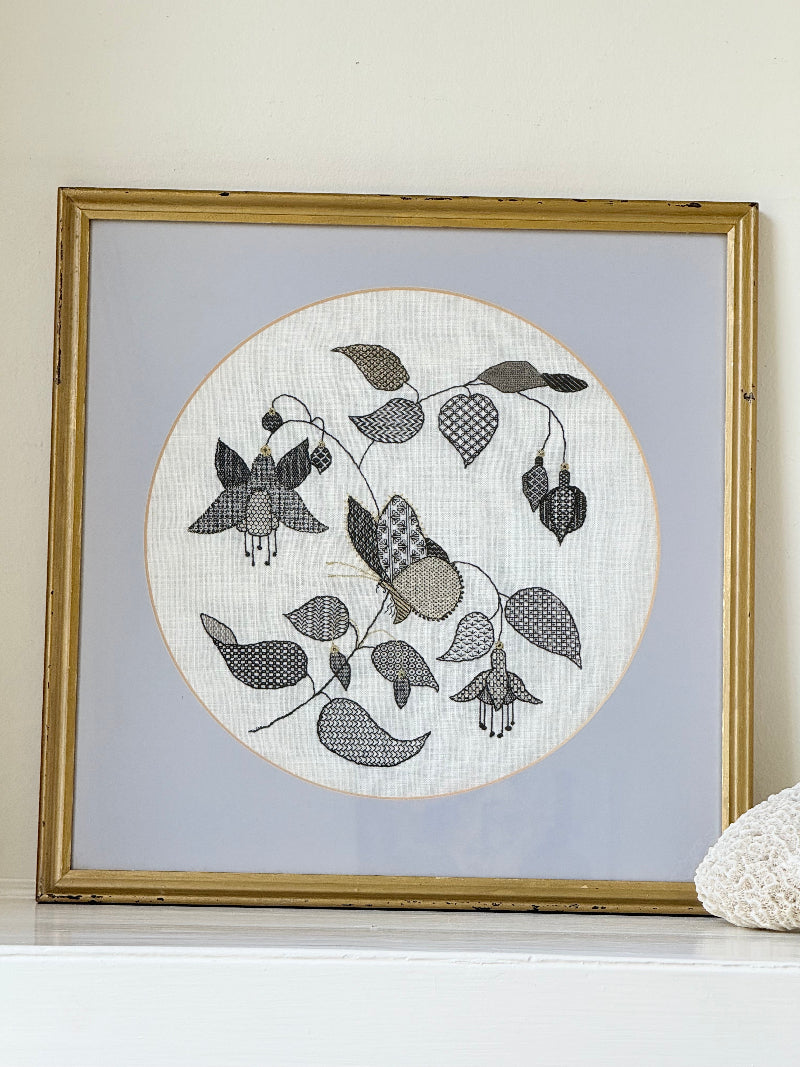 Framed circular hand embroidery of birds and fuchsia flowers in black and grey on white linen, set in gold frame, white surface background.