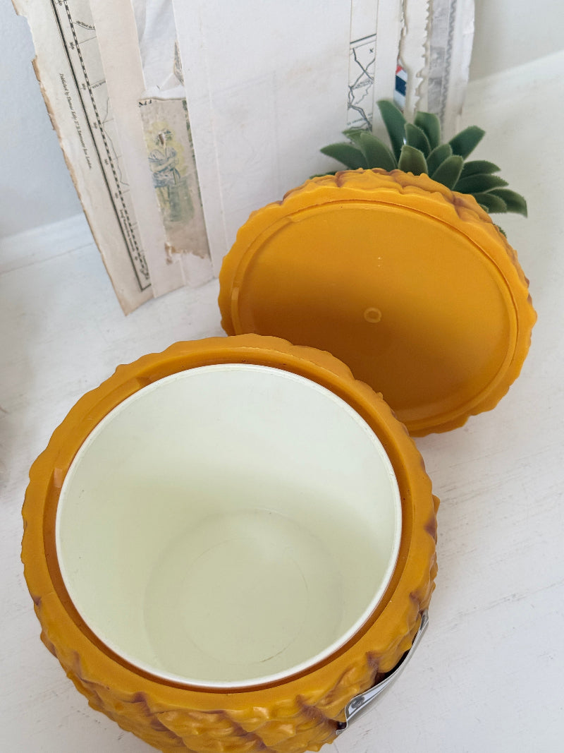 A pineapple-shaped container with a textured yellow-orange exterior and a cream-colored interior, shown with its lid propped behind it on a light surface.