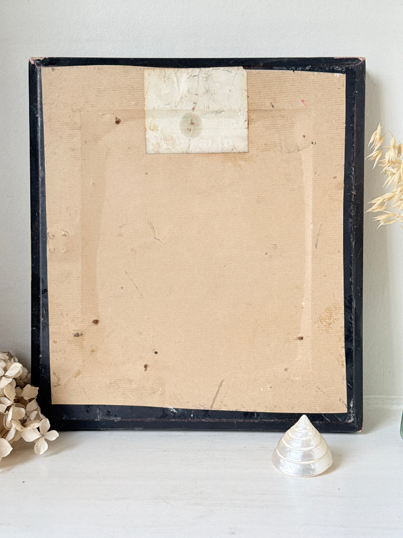 The back of a vintage black wooden picture frame leaning against a white wall on a white surface, with a beige backing board attached and dried flowers and a small shell prop around it.