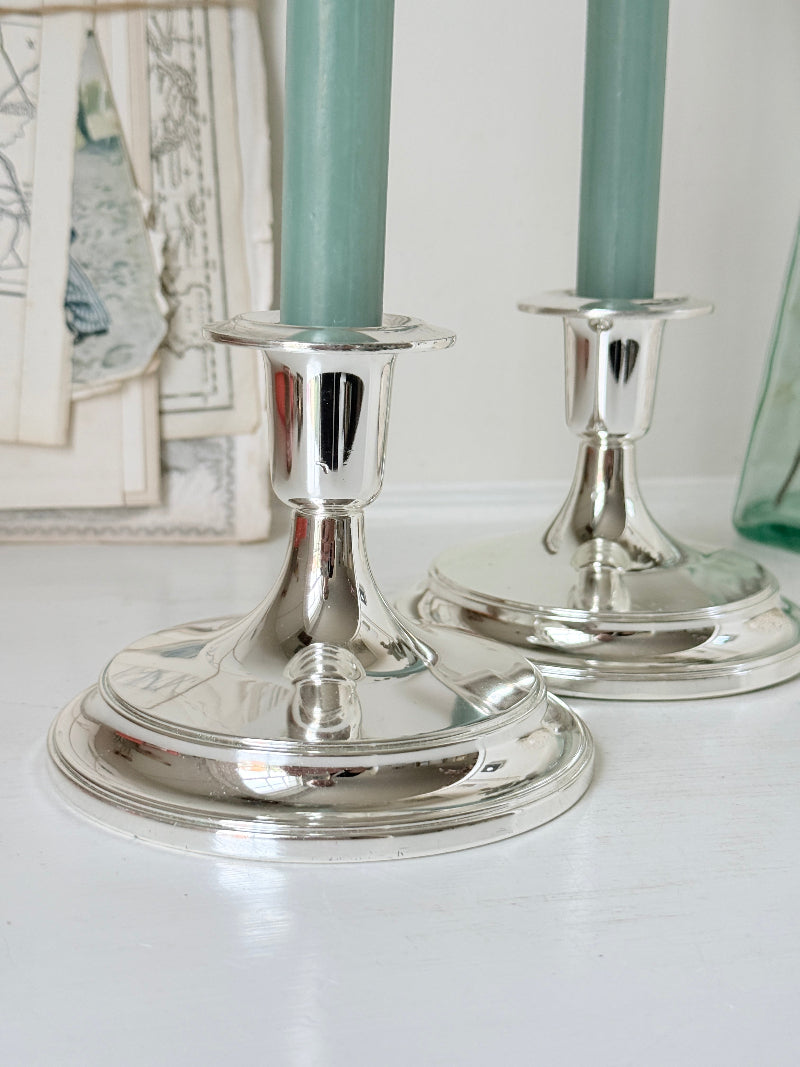 Pair of silver tone candlesticks with wide round stepped bases holding green candles, photographed on a white surface