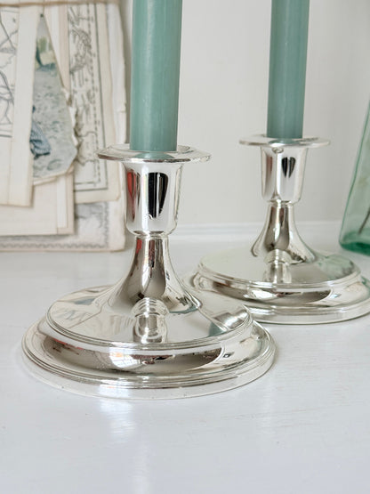 Pair of silver tone candlesticks with wide round stepped bases holding green candles, photographed on a white surface
