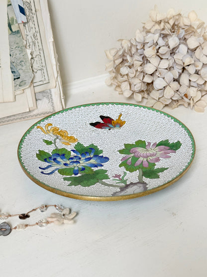 A round decorative ceramic tray with a white textured glaze, featuring hand-painted colorful flowers (blue, pink, yellow), green leaves, a butterfly, and a gold rim, styled on a white surface with dried flowers and a jewelry strand in the background.
