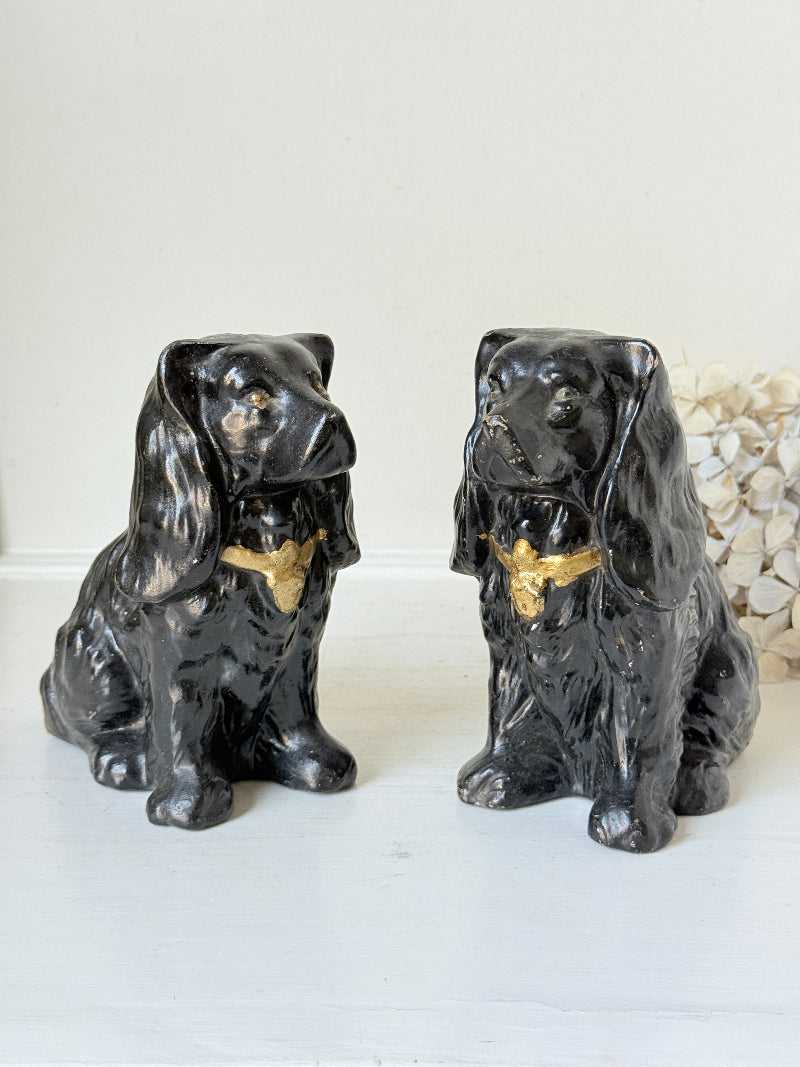 A pair of glossy black ceramic dog figurines with gold bow ties, displayed side-by-side on a white surface against a light off-white background.