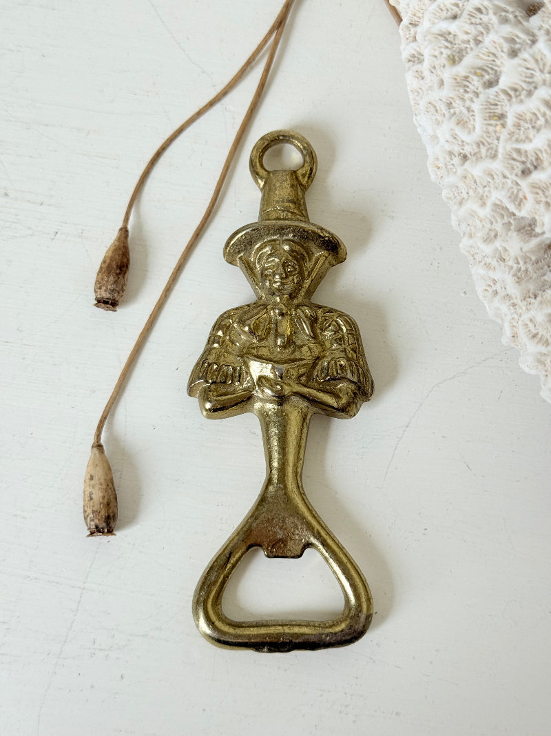 Vintage brass figure bottle opener photographed on a white surface with dried foliage.