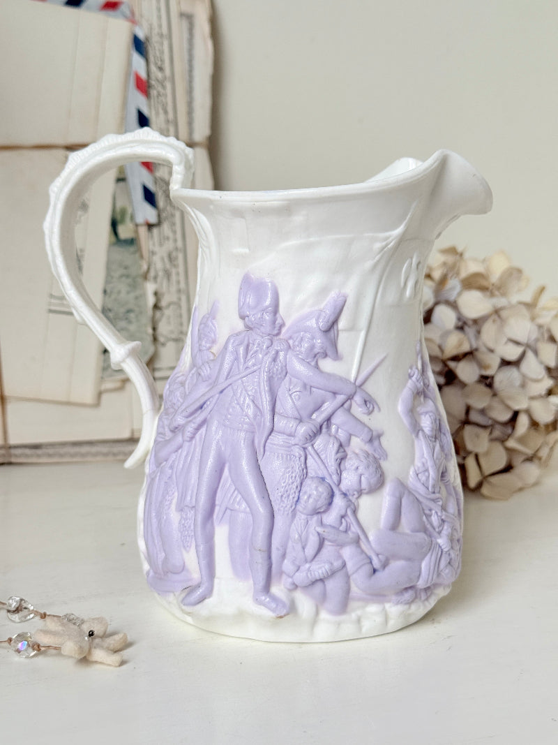 A white ceramic pitcher with a pale lavender embossed relief design depicting a historical battle scene, styled on a tabletop with vintage books and dried flowers in the background.