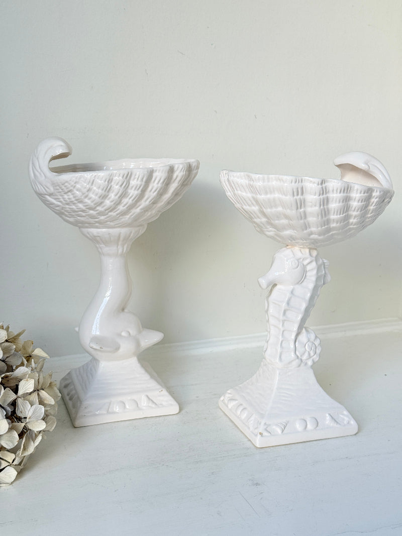 Two white ceramic nautical-themed candle holders shaped like a seashell and a seahorse, displayed on a white surface against a light wall.