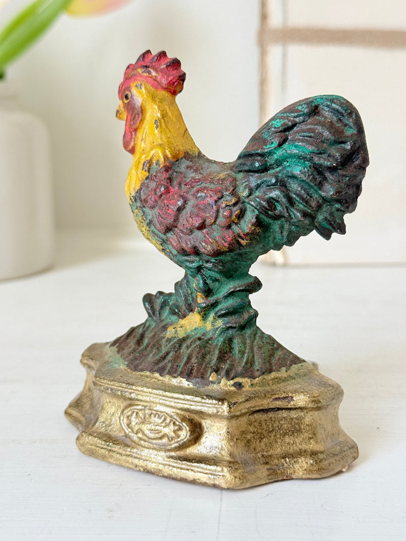 A vintage cast iron rooster figurine with a distressed multicolor finish, featuring a yellow neck, red comb, and green and purple textured feathers, standing on a gold-colored base.