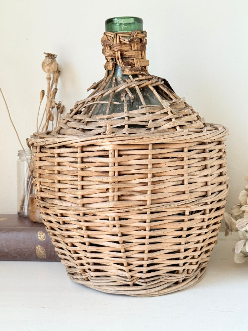 antique French demi john olive oil jar in wicker basket with green glass bottle and painted metal label, displayed on white surface