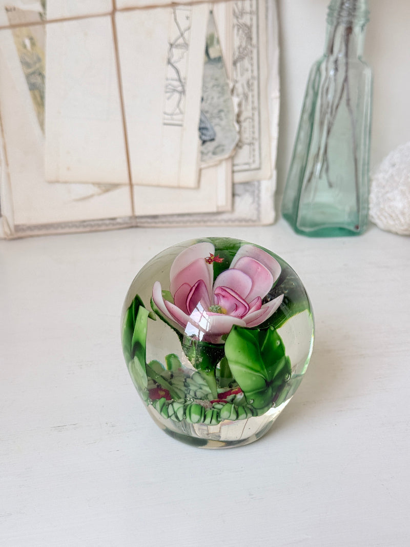 Large clear glass paperweight with a pink flower and green leaves inside, photographed on a white surface