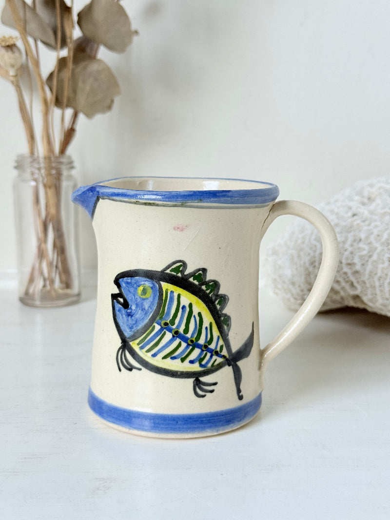 Vintage studio pottery jug with hand painted blue and green fish design on cream clay, photographed on a white surface.