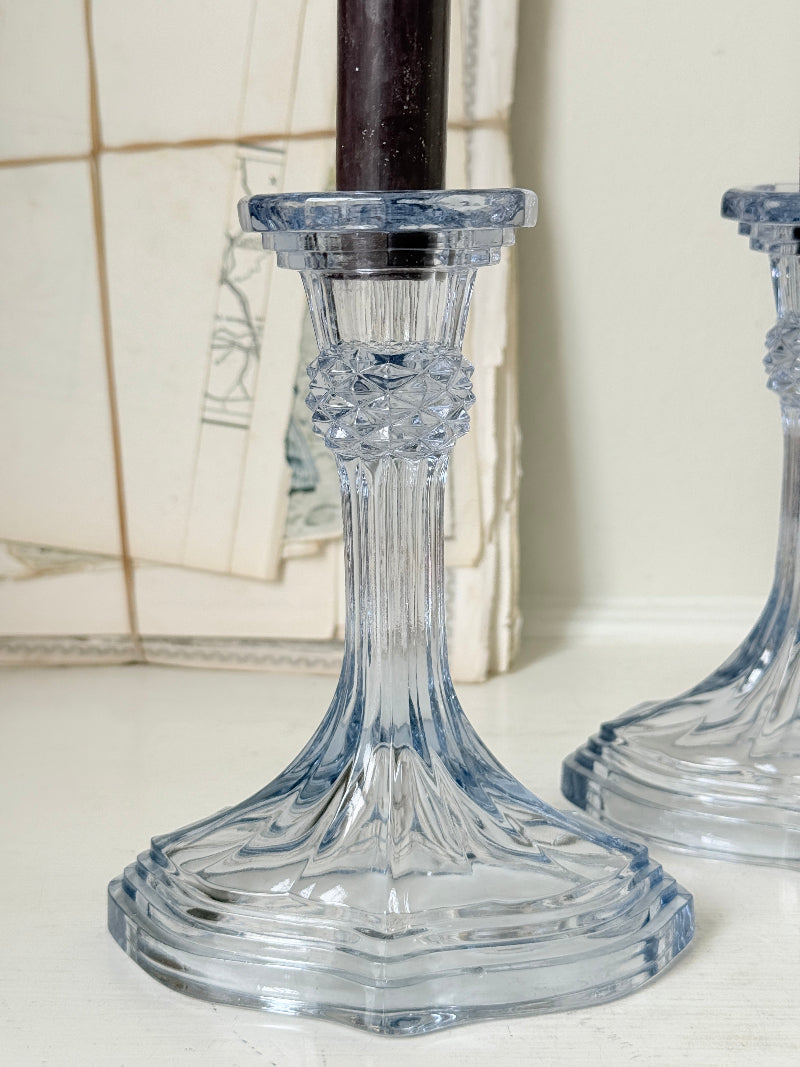 A close-up of a clear cut-glass candlestick holder with a dark brown candle inserted, set against a softly blurred background with another matching holder partially visible on the right.