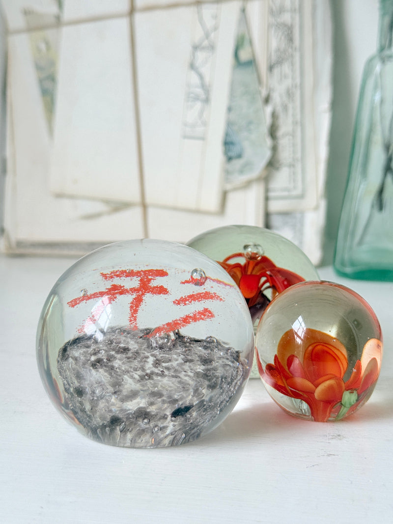 Trio of orange flower glass paperweights with green stems and controlled bubbles, photographed on a white surface