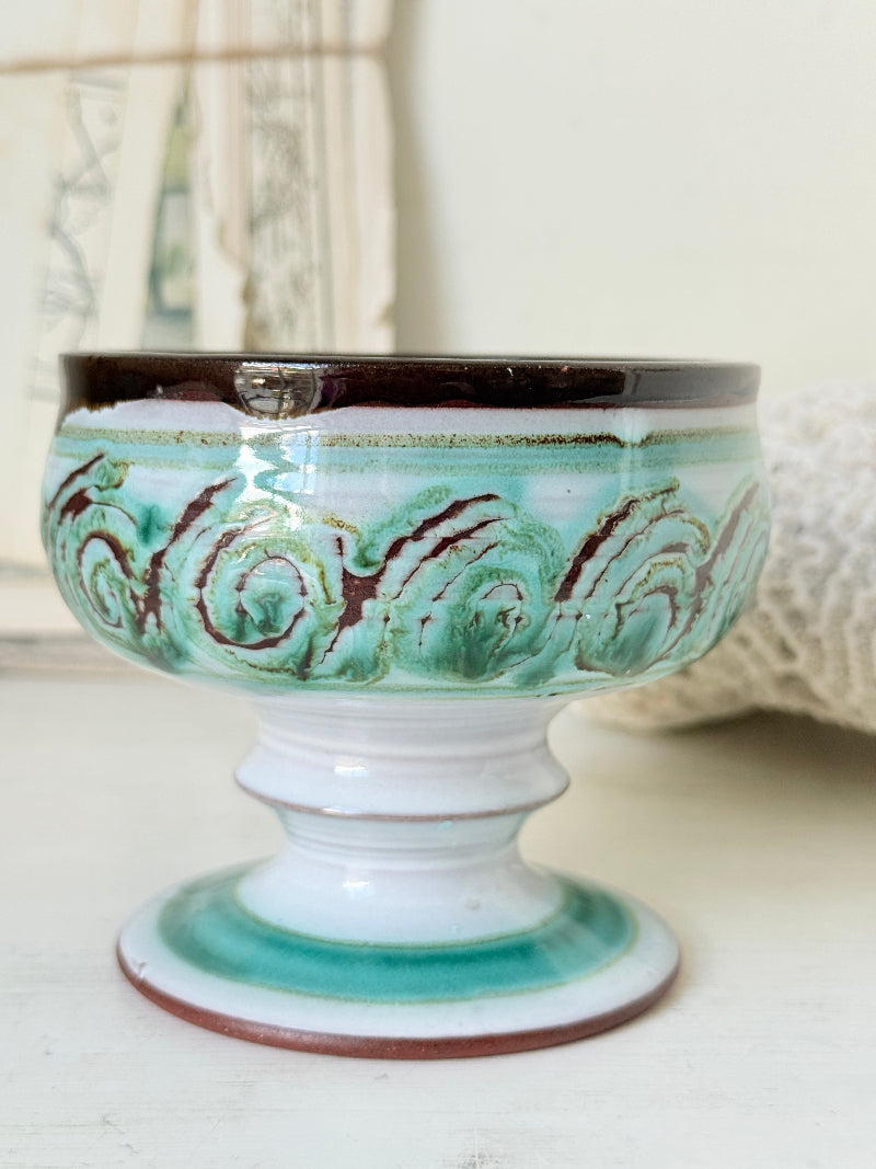 Hand thrown studio pottery footed bowl with green and brown swirling decoration photographed on a white surface