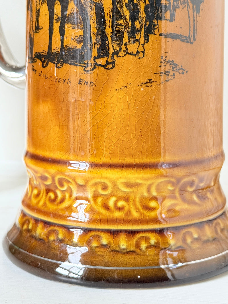 Antique Ridgways stoneware tall jug photographed on a white surface in amber treacle glaze with black transfer scenes and silver tone rim