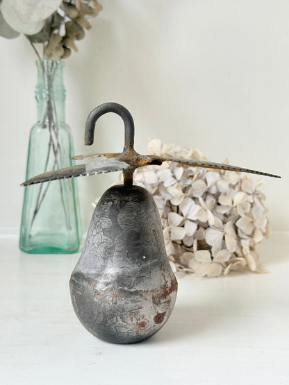 A rustic decorative metal object shaped like a pear with a curved hook on top and two long serrated blades extending from the sides, displayed on a white surface with dried flowers and a glass vase in the background.