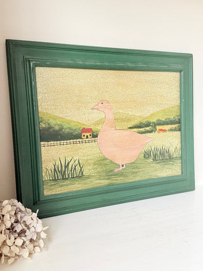 A framed folk art print featuring a pink goose in a green field, displayed on a white surface against a light beige wall with a decorative dried flower in the corner.