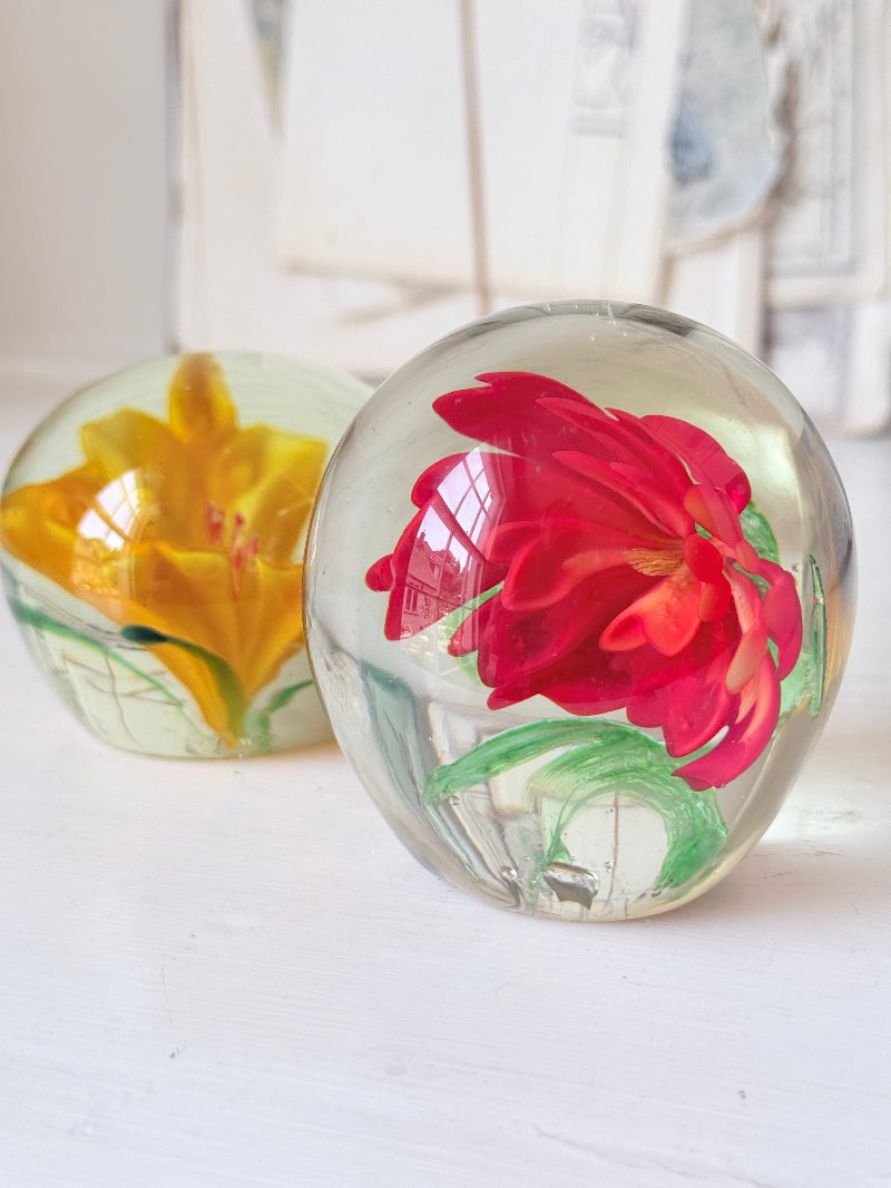 Pair of orange and yellow flower glass paperweights with green leaves, photographed on a white surface