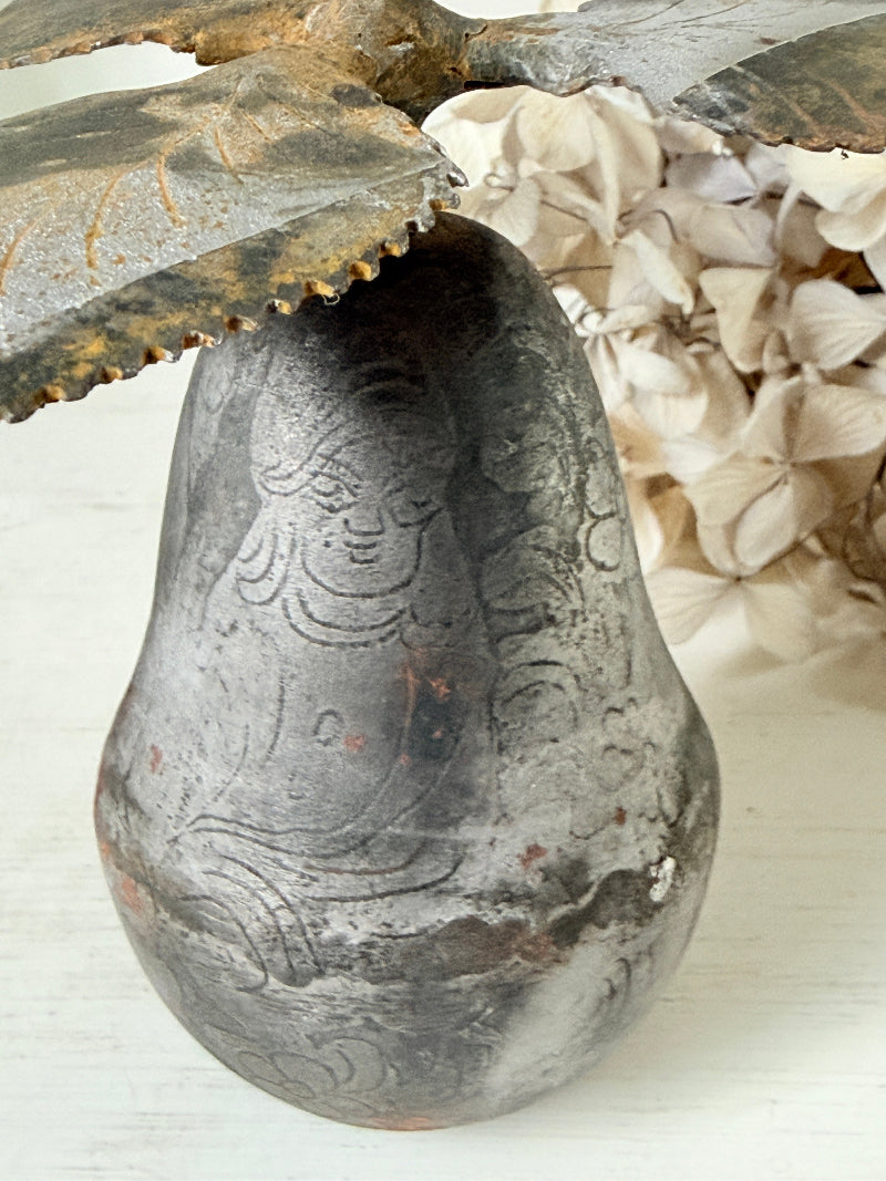 A rustic carved pear-shaped ceramic vase with a weathered gray and brown finish, styled with dried leaves and cream-colored flowers on a white surface.