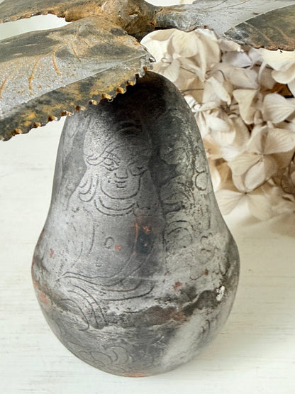 A rustic carved pear-shaped ceramic vase with a weathered gray and brown finish, styled with dried leaves and cream-colored flowers on a white surface.